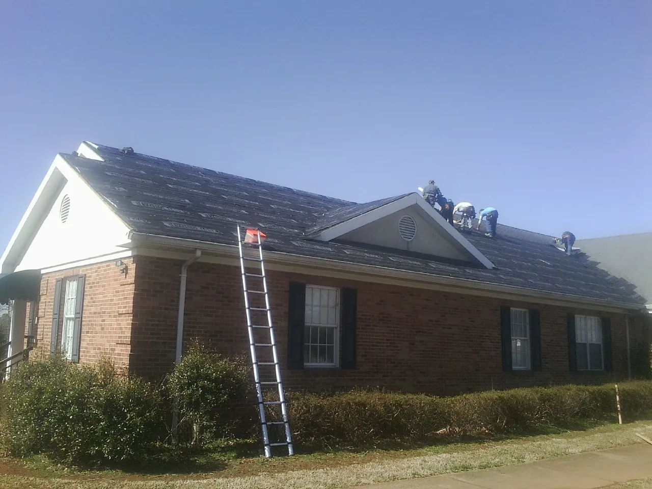 Crew tearing shingles off a brick ranch roof with a ladder set — Metal Roof Repair work by our crew in Fate