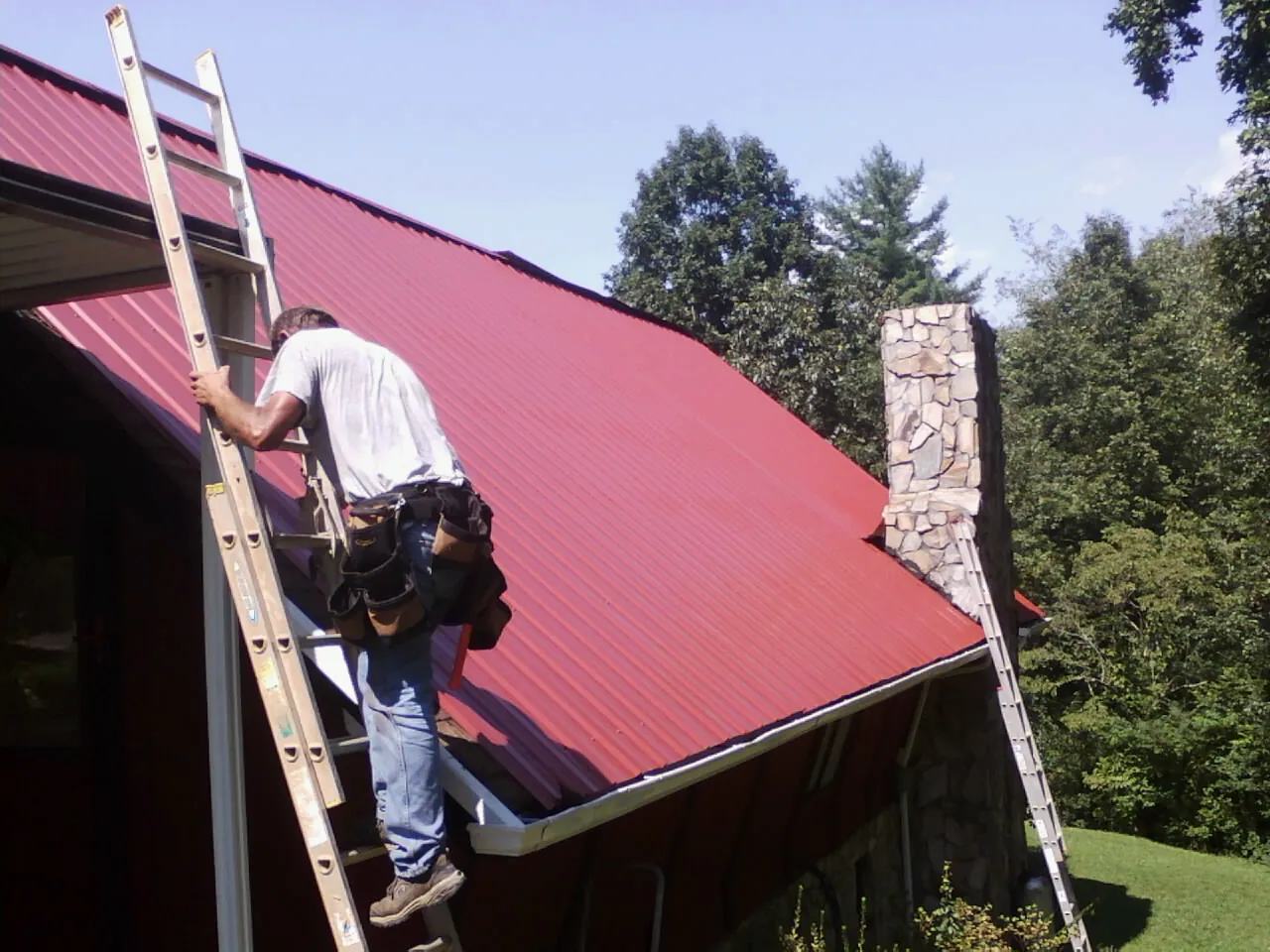 Roofer on a ladder beside a red metal roof and stone chimney — Soffit & Fascia Repair by Roofing Brothers in Fate